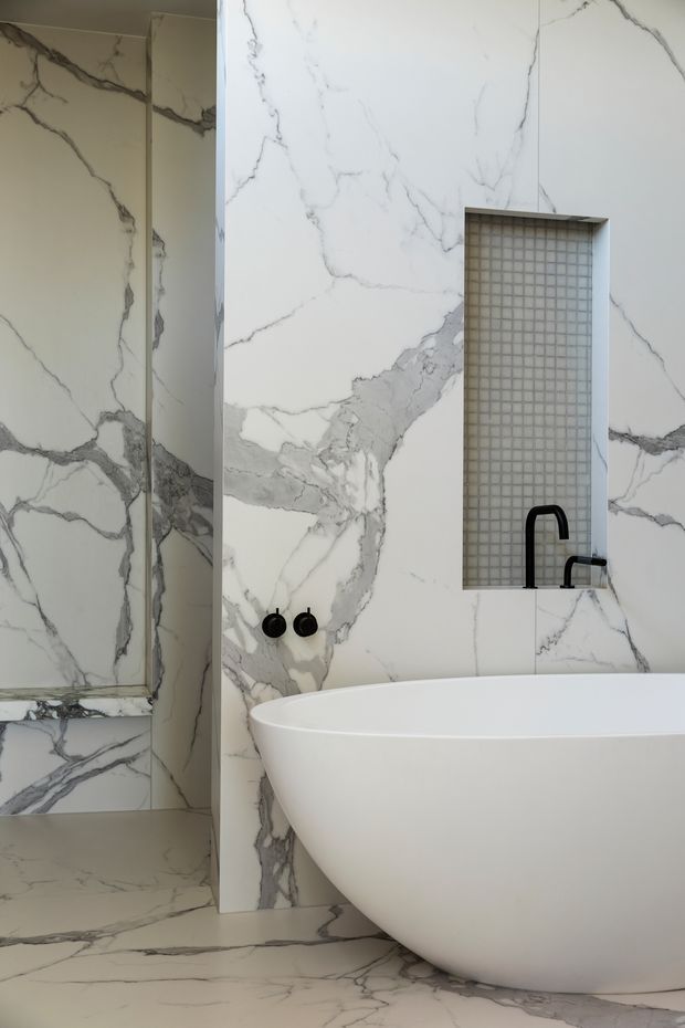 A refined contemporary bathroom clad extensively in dramatic white marble-look porcelain or stone with bold grey veining. The image shows a freestanding white bathtub in the foreground, a walk-in shower area to the left, a recessed tiled niche with black wall-mounted tapware, and minimalist black controls. The space has a clean, luxurious, monochromatic appearance.