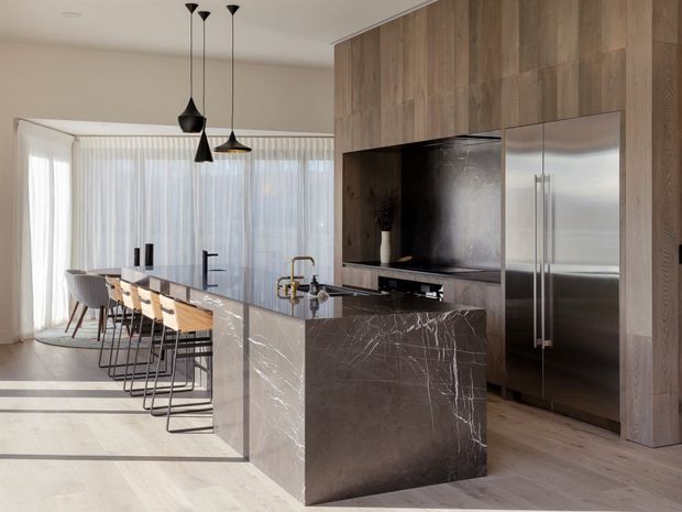 A spacious contemporary kitchen with a large dark stone island, integrated sink and tapware, timber-look full-height cabinetry, and a stainless-steel refrigerator. A row of bar stools faces the island, with sculptural pendant lights overhead and sheer floor-to-ceiling curtains enclosing a bright dining or living area.