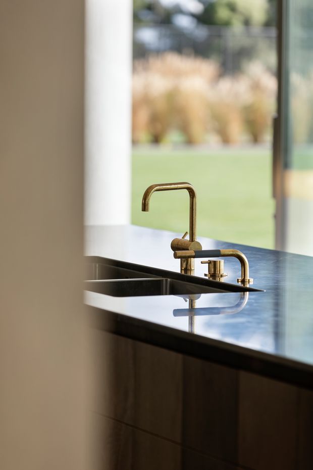 Close-up view of a contemporary kitchen island with a dark reflective stone benchtop, integrated double sink, and sculptural brass/gold-toned tapware. Dark timber cabinetry is visible below, with large glazing opening to a landscaped outdoor area.