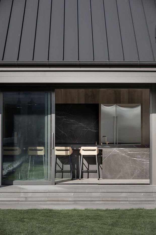 Contemporary exterior view of a compact outdoor kitchen integrated into a dark-clad building facade. The kitchen features dark marble-look surfaces, wood-toned cabinetry, stainless-steel double-door refrigeration, a sink, and two stools beneath a covered opening. A large sliding glass door is visible to the left, with a lawn in the foreground.