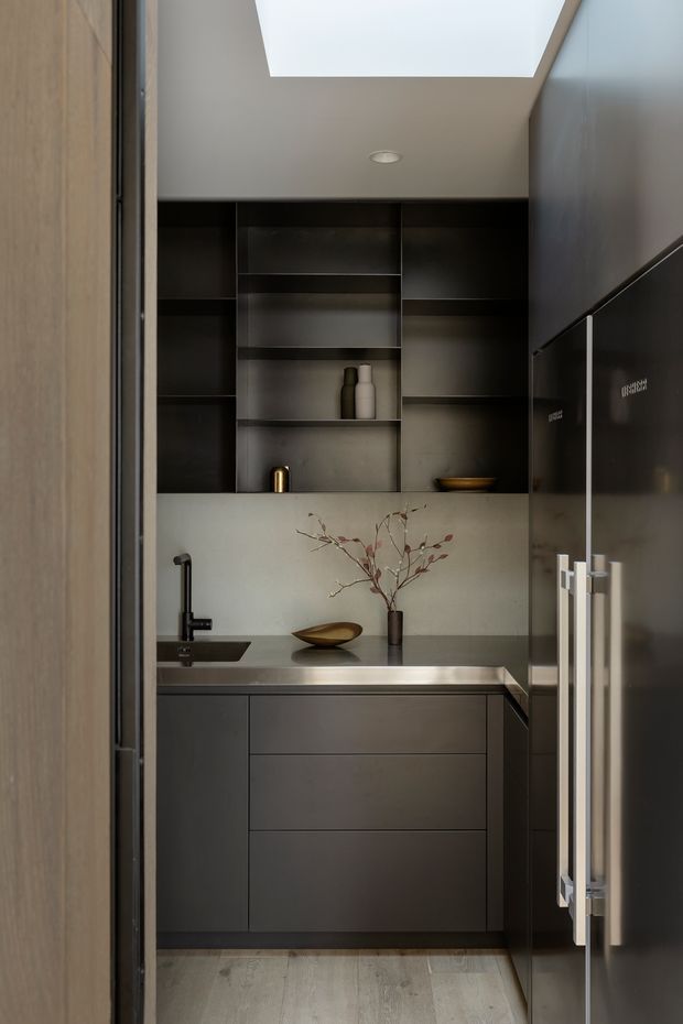 A compact, dark-toned butler’s pantry or scullery viewed through a doorway. The space features full-height dark cabinetry and open shelving, a stainless-steel benchtop, integrated appliances, a black sink and tap, a pale tiled or stone splashback, timber-look flooring, and a large skylight that brings in natural light. Minimal decorative objects and warm metallic accents add refinement.