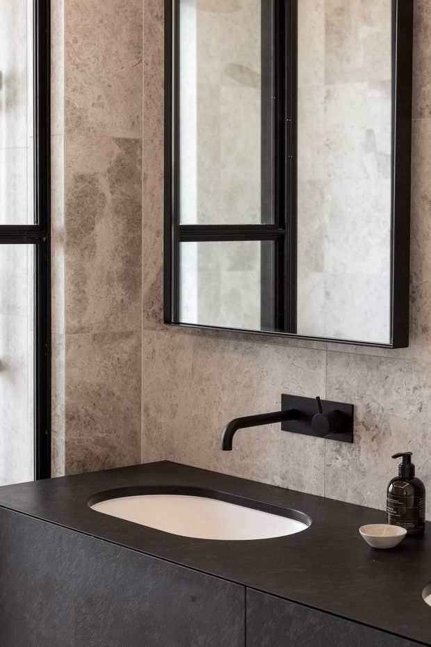 A refined contemporary bathroom vanity with a dark stone benchtop and integrated oval basin, set against large-format beige and taupe stone wall tiles. A matte black wall-mounted tap and black-framed mirrored cabinet reinforce the monochromatic, high-contrast scheme. A second basin is partially visible at the right edge, and narrow black-framed windows bring in natural light.