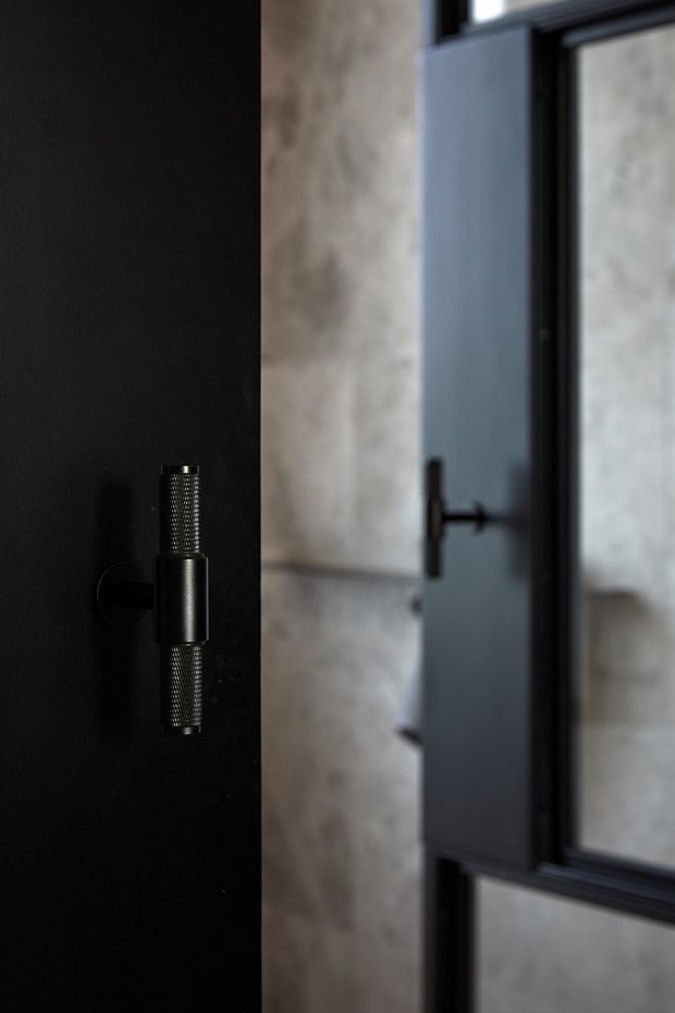 Close-up architectural detail of a dark interior door with a distinctive black metal and textured cylindrical hinge or handle. A black-framed glazed door or partition is visible to the right, with a softly textured light stone or plaster wall beyond. The image has a moody, refined, high-contrast industrial-modern character.