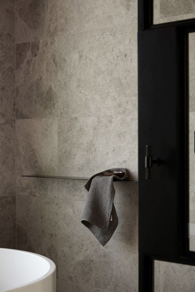 A close-up view of a refined bathroom featuring large-format grey-beige natural stone wall tiles, a slim dark metal wall shelf with a folded towel, the curved edge of a freestanding white bathtub, and a black-framed glazed door or window. The subdued palette and soft lighting create a calm, luxurious atmosphere.