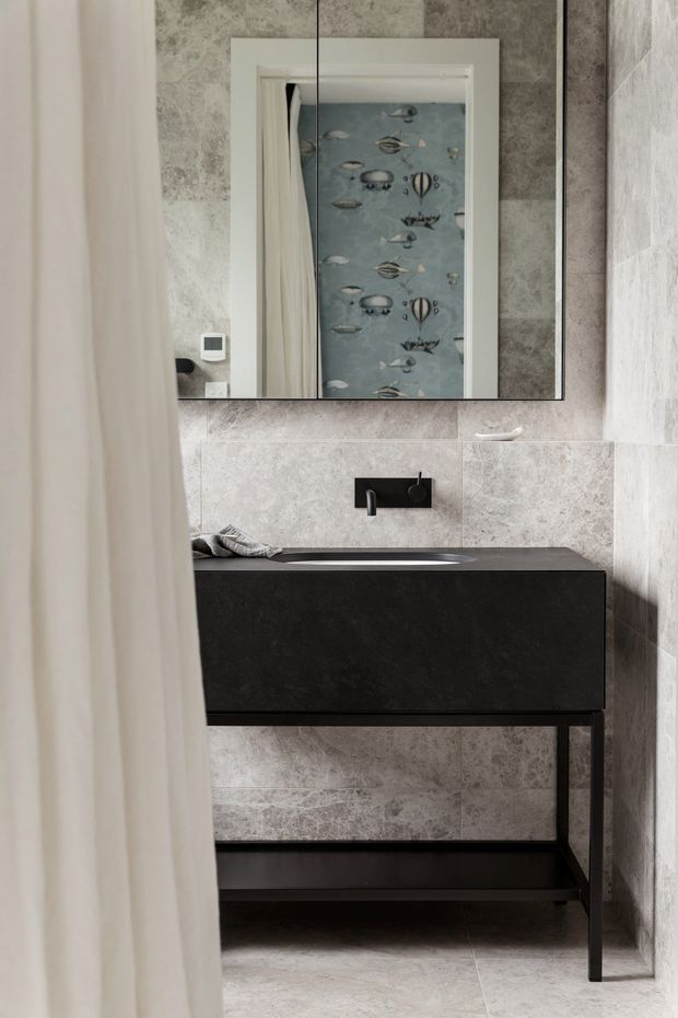 Compact contemporary bathroom viewed partly through a light curtain, featuring large pale grey stone-look wall and floor tiles, a black wall-mounted vanity with open lower shelf, black basin and wall-mounted tapware, and a large frameless mirror. A whimsical blue patterned textile or artwork is reflected in the mirror.