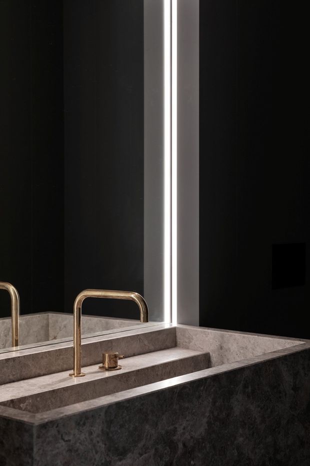 Close-up of a luxurious contemporary bathroom vanity, featuring a dark stone integrated basin and backsplash, dramatic black wall surfaces, a tall illuminated mirror, and brushed brass tapware.
