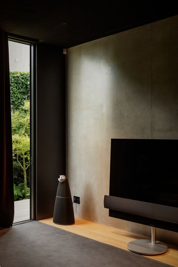 A dark, refined living room with a wall-mounted flat-screen television and slim soundbar against a textured warm-grey plaster wall. A narrow full-height glazed opening provides views to lush greenery outside, while dark curtains, a charcoal ceiling, grey carpet, and a timber floor strip create a moody, minimalist media-focused interior.