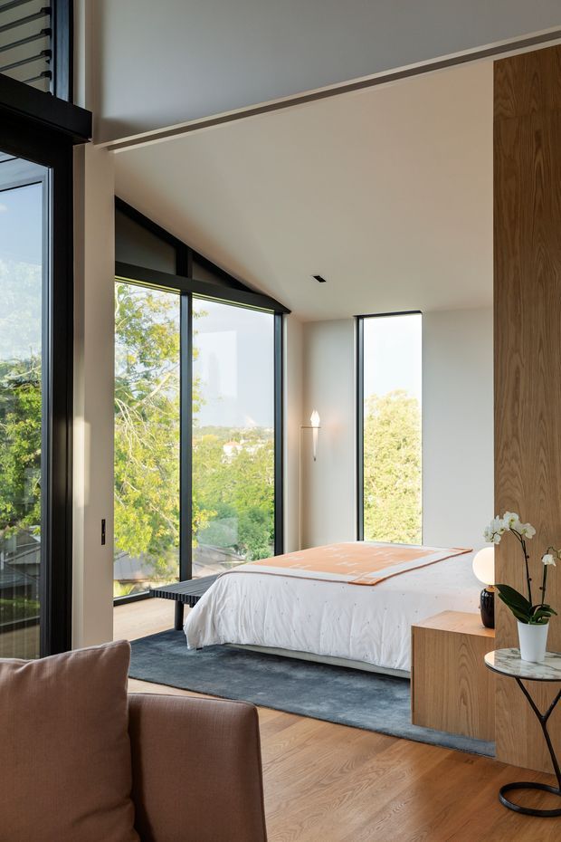 A refined contemporary bedroom viewed from an adjoining seating area, featuring a low platform bed with white bedding and a rust-coloured throw, expansive black-framed corner glazing, pale walls, warm timber flooring, and a tall timber wall panel with integrated bedside cabinetry. The room opens to leafy suburban views and has a calm, natural, light-filled atmosphere.