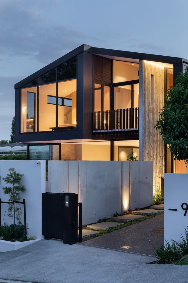 Twilight exterior view of a contemporary multi-storey residence with a strong black-framed glazed upper level, warm interior lighting, a sheltered balcony with vertical black balustrades, and a tall textured vertical timber feature wall. The property has concrete boundary walls, a dark pedestrian gate, landscaped planting, and a driveway entry.