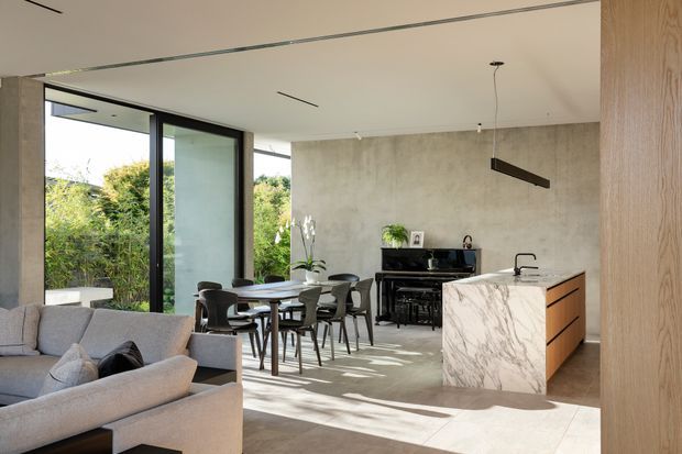 Open-plan contemporary kitchen, dining, and living area with full-height black-framed glazing opening to greenery. The space features a sculptural white marble island with timber cabinetry, a concrete-look feature wall, dark dining furniture, a grey sectional sofa, and a linear pendant light. Natural light creates a bright, refined interior with strong indoor-outdoor connection.