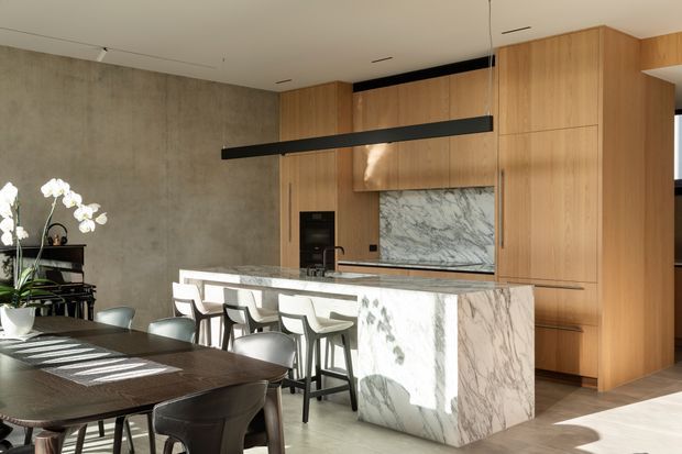 A refined open-plan kitchen and dining area with a large veined stone island and integrated waterfall end panels, surrounded by warm timber cabinetry. A dark textured feature wall contrasts with the timber joinery, while a linear black pendant hangs above the island. The foreground contains a dark dining table with contemporary chairs, and natural light creates strong shadows across the pale floor and island.