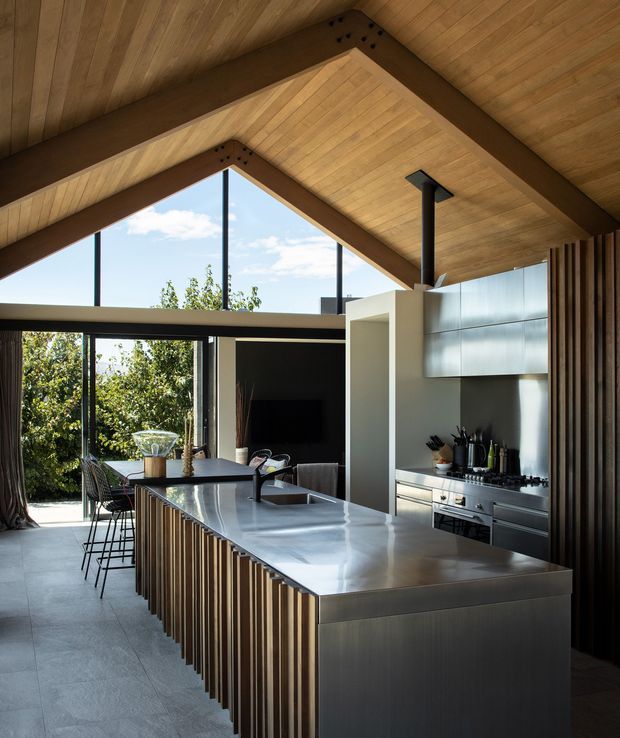 A contemporary open-plan kitchen beneath a dramatic timber-lined pitched ceiling. The room features a large island with a polished grey stone benchtop and vertically fluted timber-clad sides, integrated stainless-steel cooking appliances, streamlined pale cabinetry, dark metal framing, and full-height glazing opening toward greenery. Exposed dark timber structural beams and a high triangular clerestory create a warm, airy architectural interior.
