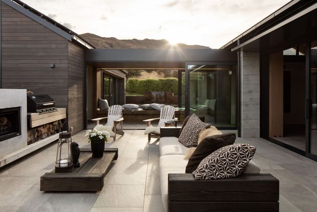 A modern, single-level courtyard entertaining area framed by dark horizontal timber-clad wings and extensive glazing. The space includes an outdoor fireplace and built-in barbecue with firewood storage, a long sectional sofa, lounge chairs, a central low fire-table or coffee table, and a covered seating nook beyond. Mountain terrain is visible in the background, while large-format pale paving and dark timber create a refined indoor-outdoor setting.