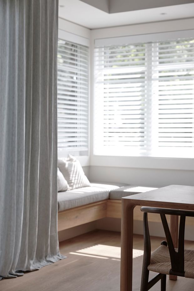 Bright, softly styled interior corner featuring a built-in timber window seat with pale upholstered cushions, white plantation shutters, sheer grey curtains, and a simple timber dining or work table with a woven-seat chair. Warm natural light falls across the timber floor and furniture, creating a calm, understated space.