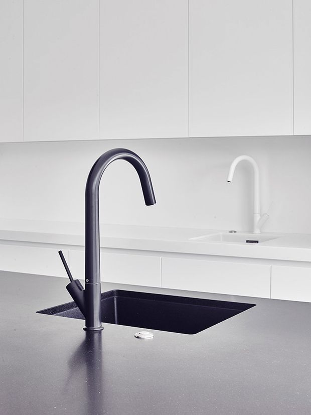 Close-up of a minimalist kitchen worktop featuring a large matte dark-grey island benchtop, integrated black sink, and tall curved dark tap. White handleless cabinetry and a secondary white sink and tap are visible in the background, creating a clean monochromatic contemporary setting.