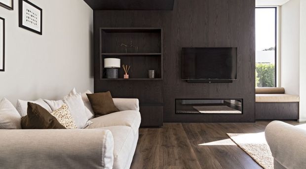 Contemporary living room with a large cream sectional sofa, dark timber feature wall, built-in shelving, wall-mounted television, and recessed linear fireplace. A tall window seat brings in natural light, while framed artwork and neutral furnishings create a warm, refined interior.