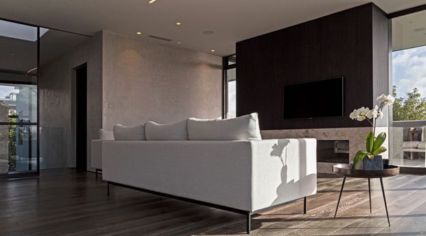 A refined contemporary living room with a large light-grey sectional sofa facing a wall-mounted television set into a dark timber feature wall. The room has dark timber flooring, textured neutral walls, full-height glazing, a linear fireplace or media joinery below the television, and a slim black-framed coffee table with an orchid. Strong natural light creates a calm, high-contrast atmosphere.