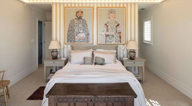 A refined bedroom with a centered upholstered bed, pale bedding, matching bedside tables and table lamps, and a dark carved wooden bench at the foot of the bed. The feature wall has vertical cream-and-white striped wallpaper with two framed decorative artworks, while the remaining walls are neutral grey with white trim. A window provides natural light, and a doorway opens to the side.