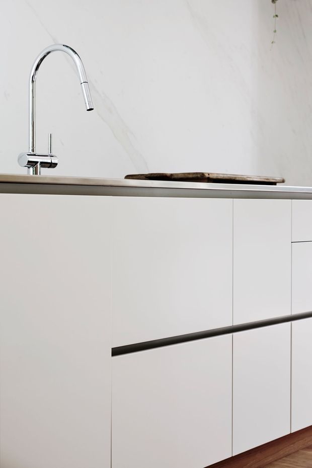 Close-up view of a minimalist kitchen with handleless white cabinetry, a slim metallic benchtop edge, a polished chrome gooseneck tap, and a pale veined stone backsplash. A dark timber cutting board rests on the benchtop, and timber flooring is visible at the base.