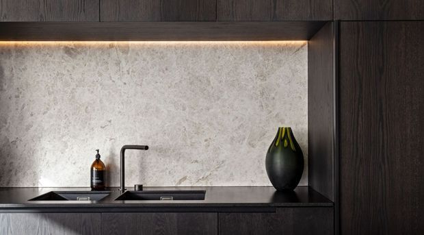 A refined contemporary kitchen vignette featuring dark timber cabinetry, a dark stone benchtop, a pale textured stone backsplash, an undermount sink with a matte black gooseneck tap, integrated warm under-cabinet lighting, and a black decorative vase.
