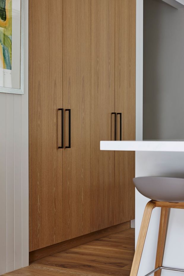 Close-up interior view of a contemporary built-in timber wardrobe or storage cabinet with vertical wood-grain doors and slim black pull handles. The cabinet sits against a light vertically panelled wall and warm timber flooring. A white island or counter edge and a modern stool with a pale upholstered seat are visible in the foreground, while a grey recessed area appears to the right.