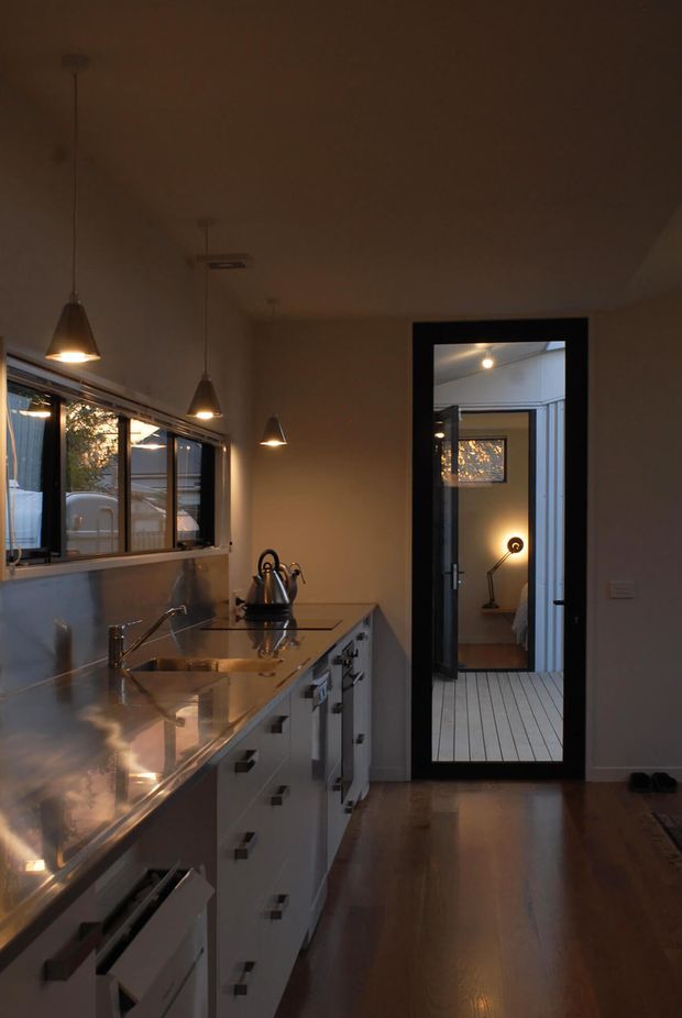 A warm, dimly lit contemporary kitchen viewed along a long run of white cabinetry and a reflective stainless-steel benchtop. Multiple pendant lights hang above the work surface, with a stainless sink and tap beneath a row of windows. A black-framed glazed doorway opens toward a bedroom or living space beyond, and polished timber flooring continues through the room.