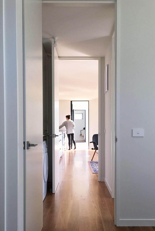 A bright, narrow residential hallway viewed through an open doorway, with continuous timber flooring, white walls and trim, simple track lighting, and multiple aligned doorways leading toward a light-filled room. A person stands near a compact workspace or kitchen area in the distance, with a chair, mirror, and small patterned rug visible.