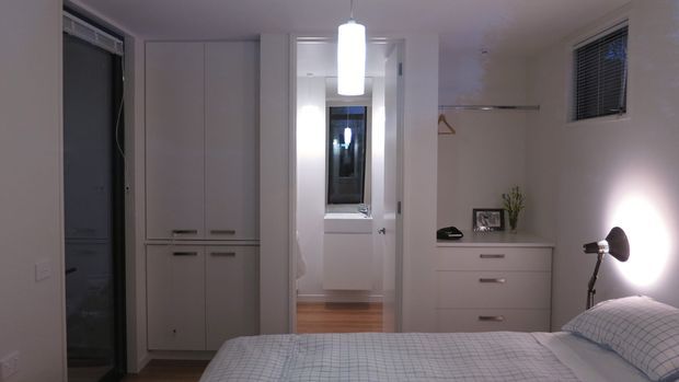 A compact, softly lit bedroom viewed from beside the bed. The room features a white built-in wardrobe, a low white chest of drawers with simple metal handles, a small framed photograph and plant, a floor lamp, and a neatly made bed with a pale checked cover. An open doorway leads to a bright adjoining area with timber flooring, while a high horizontal window has venetian blinds. The overall fit-out is simple, clean, and contemporary.