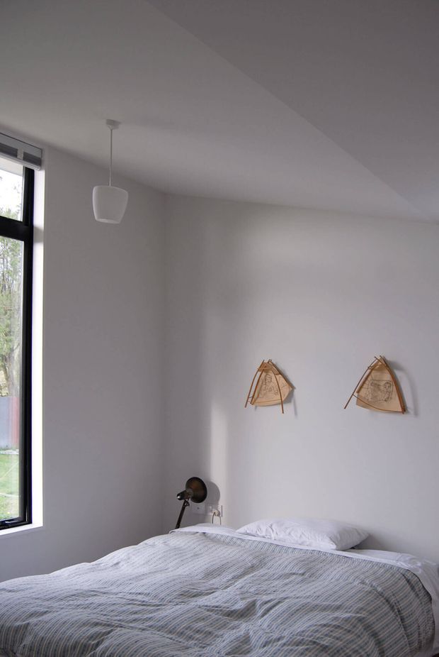 A minimalist bedroom with a neatly made bed, white textured bedding, plain white walls and ceiling, and a tall black-framed window bringing in natural light. A small dark bedside lamp sits beside the bed, while two triangular wooden wall artworks provide subtle decorative detail. The room has a simple, calm and uncluttered appearance.