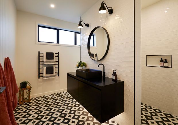 A contemporary bathroom with a floating dark vanity, rectangular vessel basin, round framed mirror, wall-mounted black sconces, white tiled walls, and a bold geometric black-and-white patterned floor. A heated towel rail, recessed shower niche, and frameless glass shower partition are visible.