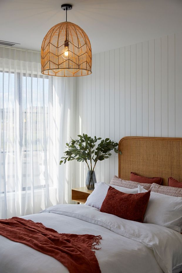 Bright, airy bedroom featuring a large bed with white bedding, rust-red cushions and throw, a woven rattan/cane headboard, white vertical wall panelling, sheer floor-to-ceiling curtains, a timber bedside table, leafy greenery, and a prominent woven pendant light. The styling is warm and natural with coastal and contemporary influences.