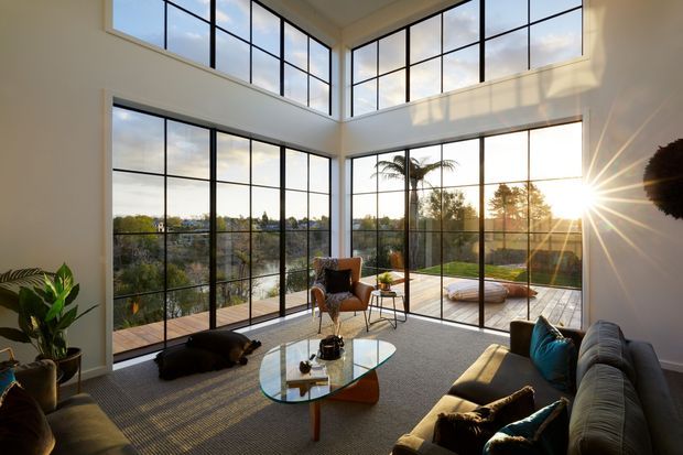 A bright, double-height contemporary living room with expansive black-framed floor-to-ceiling glazing on two sides, opening to a timber deck and landscaped garden. The room includes a large sectional sofa, armchair, glass coffee table, indoor plants and a textured neutral rug, with warm late-afternoon sunlight streaming across the space.