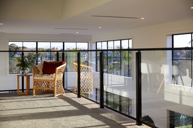 Bright modern upper-level interior or landing with expansive black-framed glazing and glass balustrades overlooking a lower level. The space has light carpet, white walls and ceiling, strong natural light, and a rattan lounge chair with a small timber side table. Reflections and views through the glazing reveal surrounding trees and neighbouring buildings.
