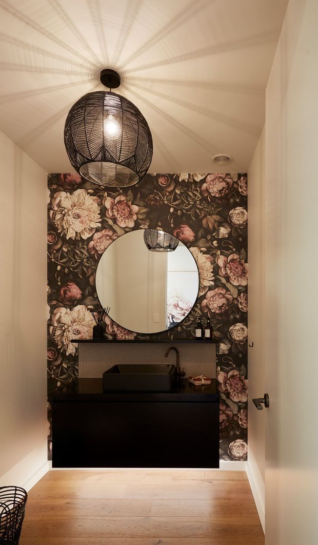 A compact powder room viewed through an open doorway, featuring a black floating vanity with integrated basin, round wall mirror, dark floral botanical wallpaper, and a large woven pendant light. Warm timber flooring and soft cream walls create a moody, richly patterned interior.