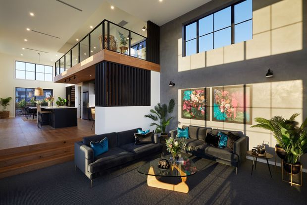 A spacious contemporary open-plan living area with a double-height ceiling, expansive black-framed windows, and a glass-balustraded mezzanine overlooking the room. The space includes a dark sectional seating arrangement, sculptural glass coffee table, indoor plants, colourful artwork, timber flooring and stairs, and a modern kitchen with a central island in the background. Strong natural light creates geometric shadows across the feature wall.