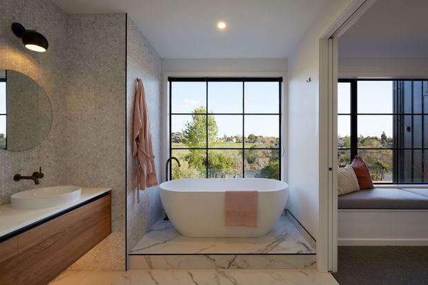 Bright contemporary bathroom featuring a freestanding white bathtub set on a raised marble platform beside large black-framed windows. The room includes a timber floating vanity with white basin, a textured small-format mosaic tile feature wall, black tapware and lighting, and a glass-panelled opening to a carpeted bedroom or sitting area.