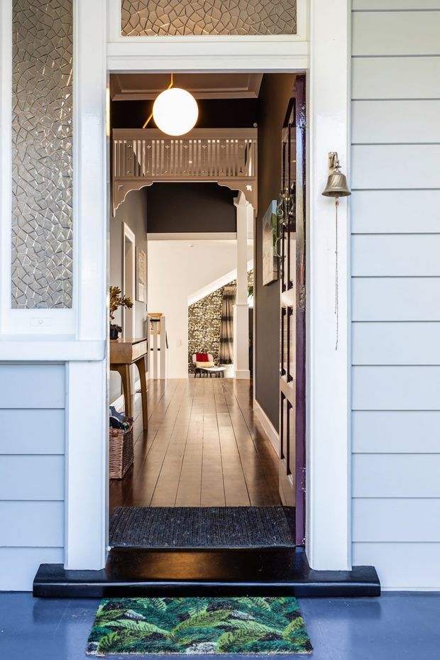 View from outside through an open front door into a richly detailed residential entry hall. The exterior has pale blue horizontal weatherboard cladding, white trim, and textured leadlight-style glass panels. Inside, a long timber-floored hallway features dark painted walls, decorative timber architraves and fretwork, a raised balustrade or mezzanine detail, framed wall art, a console table, and a glowing spherical pendant light. A patterned doormat sits on the blue-painted exterior threshold.