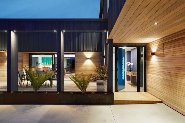 A contemporary courtyard-style residential exterior at dusk, featuring dark vertical metal cladding and framing, warm timber-lined soffits and walls, integrated lighting, glazed sliding doors, a planted raised garden bed, and a brightly lit entry vestibule.