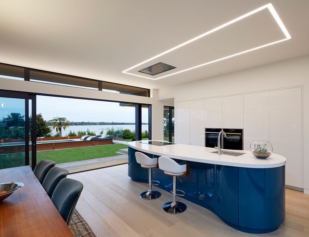 Contemporary open-plan kitchen and dining area with a sculptural curved navy-blue island, white stone benchtop, integrated appliances, and white full-height cabinetry. Large dark-framed glazing opens to a landscaped waterfront-facing outdoor area, while a linear recessed ceiling light and timber flooring complete the refined interior.