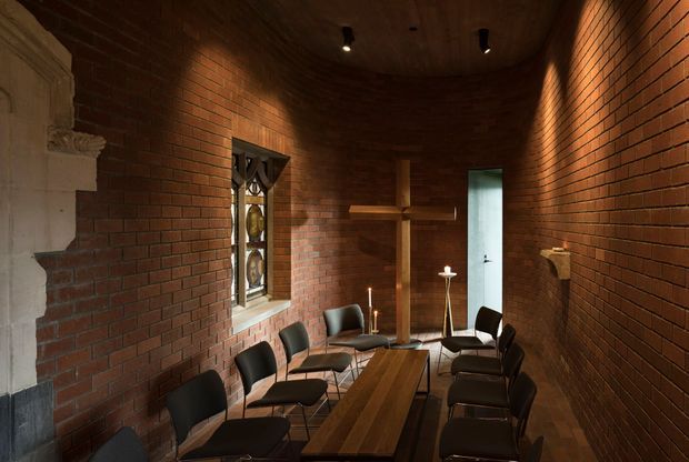 A narrow, dimly lit brick-lined interior arranged as a meeting or dining room, with a long timber table surrounded by black chairs. The space has a dark timber ceiling, exposed red brick walls, recessed spotlights, a small decorative leaded-glass window, and a tall narrow doorway at the far end. A partial pale stone or plaster architectural feature is visible at the left edge.
