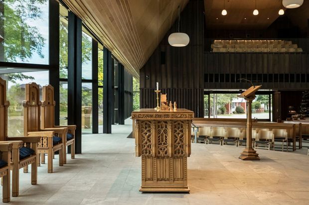A spacious contemporary chapel or church interior with a highly ornate carved timber altar or lectern in the foreground. Tall black-framed windows line one side, bringing in abundant natural light and views of greenery. The ceiling is clad in warm timber boards, while the rear wall is dark vertically ribbed timber with a raised gallery or choir area, pendant lighting, and rows of simple timber seating. Additional carved timber chairs and a lectern reinforce the ecclesiastical setting.