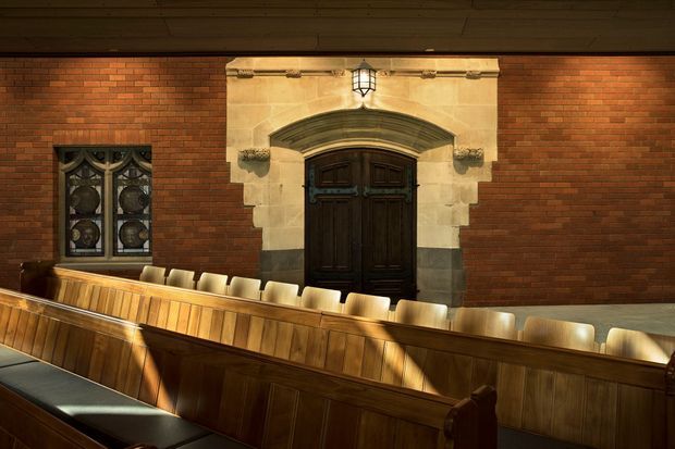 A formal wood-panelled meeting or council chamber with long tiered timber benches and upholstered seating. The room features extensive red brick walling, a prominent dark timber double door framed by an ornate pale stone architrave, a small leaded decorative window, and warm traditional lantern lighting. The architecture has a strong institutional and historic character.
