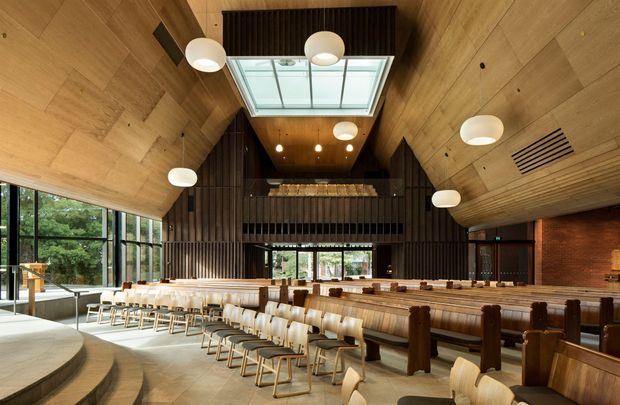 A spacious contemporary hospitality or dining interior with a dramatic timber-lined vaulted ceiling, exposed dark timber slat wall, large clerestory skylight, and extensive floor-to-ceiling glazing. Long communal timber tables and rows of light upholstered chairs surround a central bar or service counter, while curved stepped seating occupies the foreground. A brick wall and warm globe pendant lighting add texture and warmth.