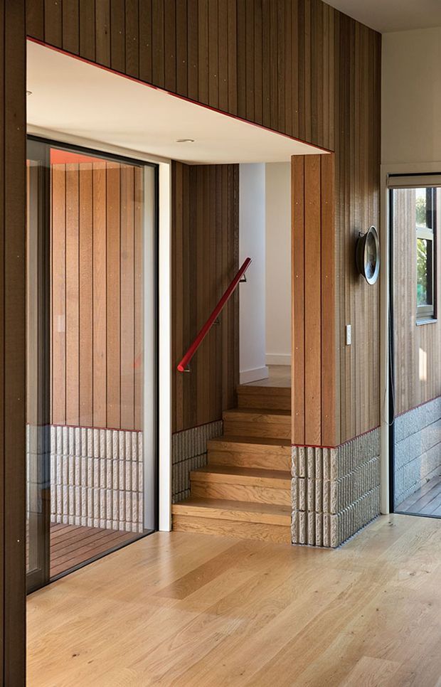 Interior view of a contemporary entry hall and stairway. Warm timber flooring and vertical timber wall panelling wrap around a compact staircase, with a distinctive red handrail, white plaster ceiling, glazed doors, and textured masonry or tile cladding at the lower walls.