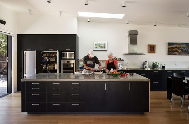 Bright, spacious contemporary kitchen with a large dark island, integrated appliances, open shelving, and a stainless-steel rangehood. Two people are preparing food at the island. White walls and ceiling, timber flooring, extensive dark cabinetry, skylight, and a dining area are visible.