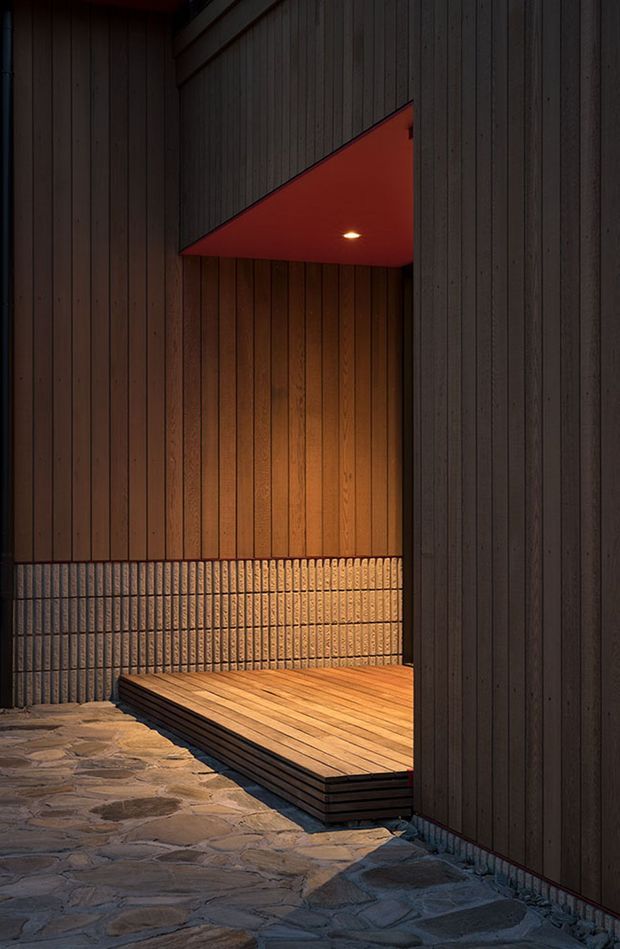 A moody architectural exterior detail showing a recessed entry or covered outdoor threshold. Dark vertical timber cladding frames a warm timber deck platform, with a red-painted soffit and recessed downlight above. Rough irregular stone paving extends across the foreground, while a textured ribbed masonry or tile wall forms the rear surface.