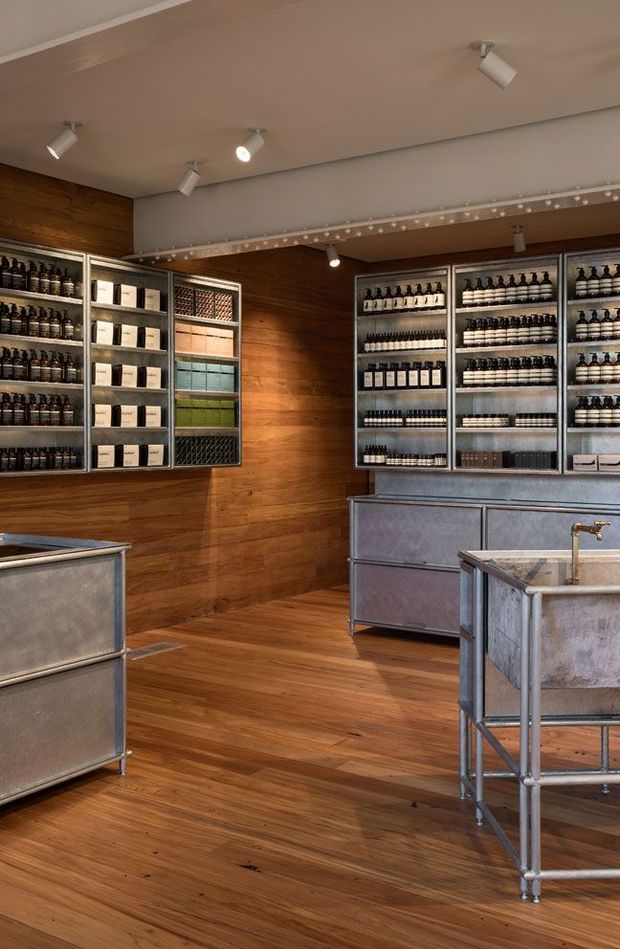 A warmly lit retail interior with floor-to-ceiling timber wall and flooring, industrial-style metal shelving densely stocked with bottles and boxed products, and large galvanized metal display counters with integrated sinks or basins. Track spotlights illuminate the merchandise, while a decorative exposed structural beam runs across the ceiling. The space has a refined industrial apothecary or specialty retail character.