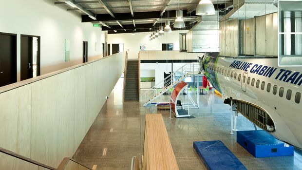 A bright, expansive commercial interior resembling an aviation training or museum facility. The image looks along an upper-level corridor with pale partition walls, polished concrete flooring, exposed ceiling services, suspended white pendant lights, and a timber bench. A large aircraft fuselage display dominates the right side, with aviation equipment and stairs visible below.