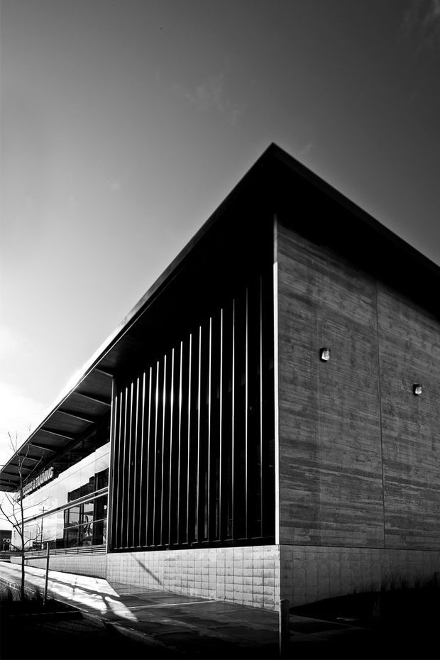 High-contrast black-and-white photograph of a contemporary building exterior. The composition emphasizes a sharply pitched gable roof, a dark vertical slatted facade, and a large textured concrete wall. A paved pedestrian edge, fencing, and glazed elements are visible along the side, with strong directional shadows creating a dramatic architectural study.