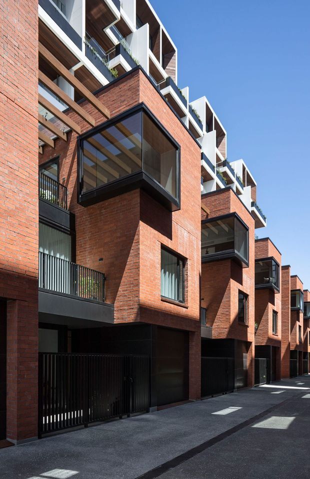 Exterior architectural photograph of a contemporary multi-storey residential development viewed from a narrow paved lane. The buildings feature prominent red brick masonry, projecting box bays with dark metal-framed glazing, black metal garage doors and gates, balconies, timber soffits, and repeated white geometric rooftop elements. Bright clear daylight emphasizes the strong vertical and angular facade composition.
